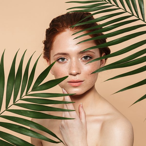 Beauty portrait of an attractive young topless redhead woman standing isolated over beige background, posing surrounded with tropical leaves